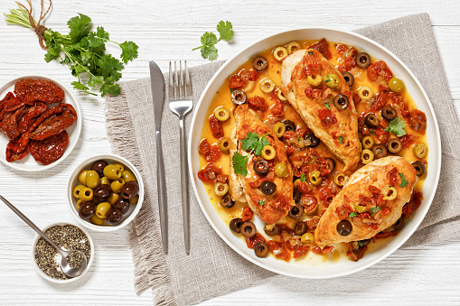 chicken breasts braised with chopped tomatoes, onion and mixed olives on white plate on textured wooden table with cutlery and ingredients, horizontal view from above, flat lay