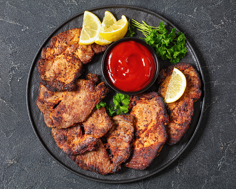 fried juicy pork steaks with ketchup, lemon slices and parsley on plate on concrete table, horizontal view from above, flat lay, close-up