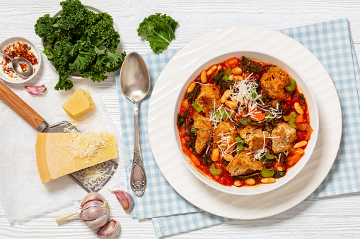 ribollita, tuscan vegetarian soup of white cannellini beans, kale, onions, carrots, celery, bread, tomatoes in white bowl on white wooden table with ingredients and spoon, flat lay