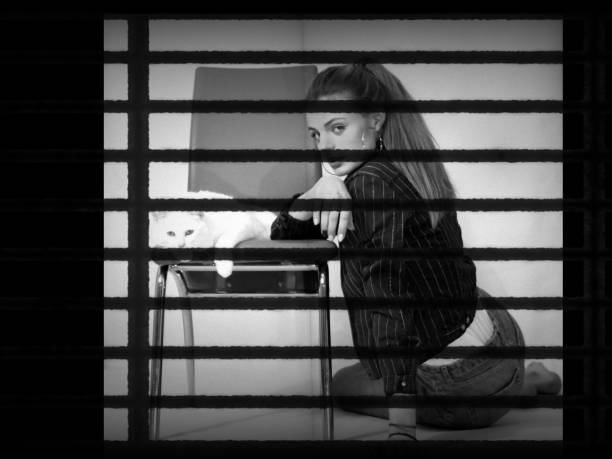 A woman sits in a pensive pose, looking out of the frame while leaning on a soft cushion by the window, bathed in contrasting shadows of elegant monochrome light.