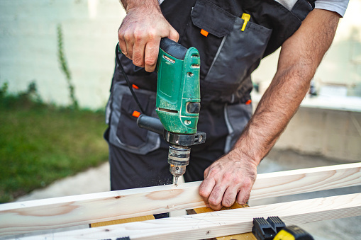 Unrecognizable male carpenter using electric drill on a wooden plank outdoors.