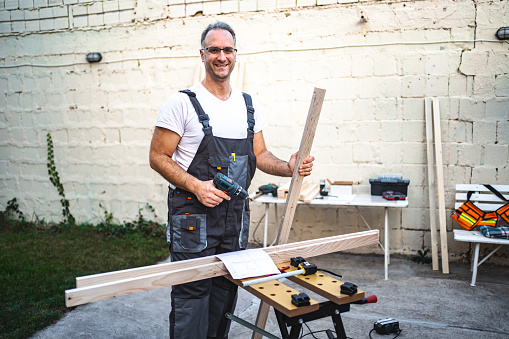 Mature male carpenter working with electrical screwdriver on a wooden plank outdoors.