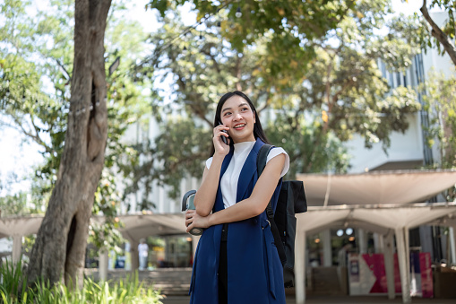 Young Asian woman stands talking on the phone in a green park, carrying a backpack. And use reusable cup to avoid damaging the environment..