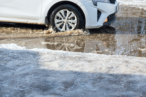 A white car drives through a deep muddy puddle of snow despite the winter thaw. Background.