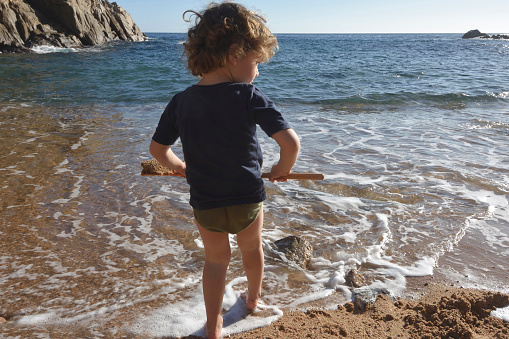 boy standing in front of the sea playing with a shovel