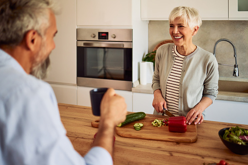 A cheerful senior adult woman enjoying a pleasant conversation with her husband while cutting vegetables and preparing a salad in the kitchen.