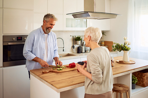 A cheerful senior adult married couple having a laugh in the kitchen while the husband is cutting vegetables and preparing a salad on a kitchen counter.