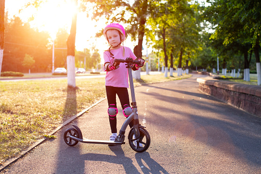a beautiful girl in a pink helmet and protective equipment stands with a scooter on an alley in the city against the backdrop of a sunny sunset in summer, sport