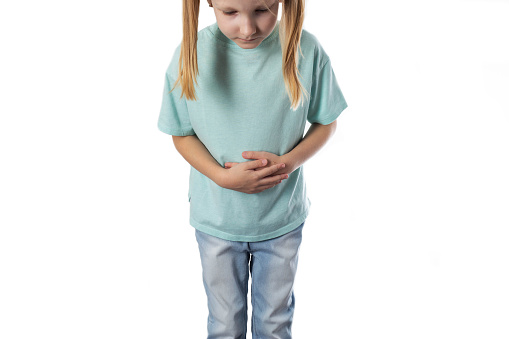 A seven-year-old blonde girl stands and holds her stomach on a white background. The concept of abdominal pain in children due to poor nutrition. Inflammation of the digestive system, dysbiosis and bloating.