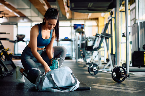 Young athletic woman packing her water bottle in sports bag after working out in a gym. Copy space.