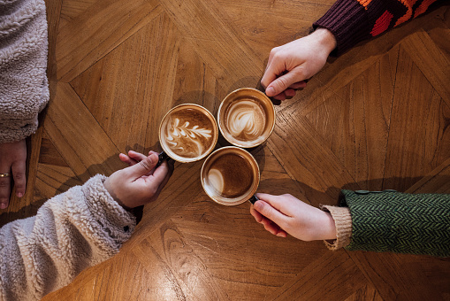 Direct above shot of friends sitting in a cafe enjoying a hot drinks with froth art. They are toasting their cups together in Tynemouth, North East of England.