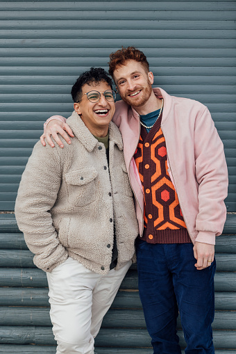 Portrait of an LGBTQI+ couple standing on a beach in Tynemouth, North East England. They are laughing with their arms around each other.