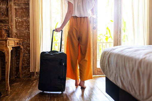 Rear view of unrecognizable woman walking out of hotel bedroom with suitcase. Vacation, trip concept.