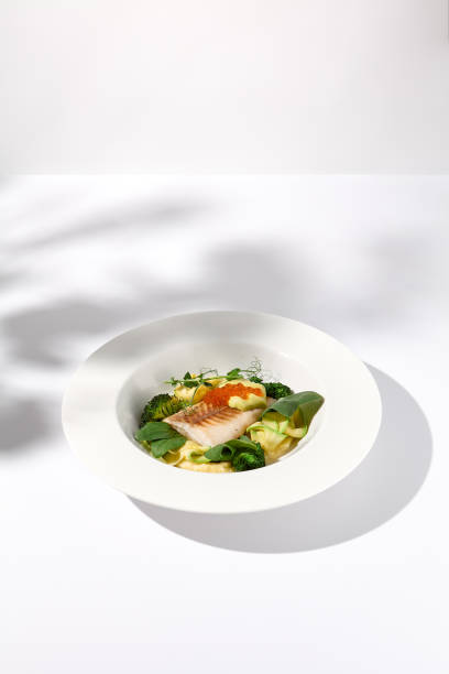 istock Cod fillet with caviar and zucchini-broccoli garnish, side view on white plate with delicate leaf shadow 2027322073