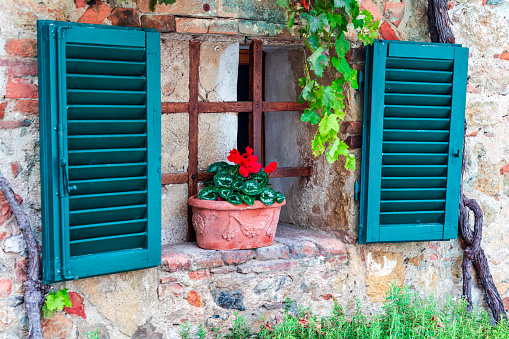 Small fairy tale style window in old stoned house with open green shutters. Clay made flower pot with red flower on stoned windowsill. Monteriggioni, Italian village
