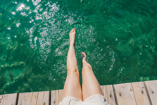 Woman relaxing by the lake on a sunny day. Feet hanging over a wooden dock above the clear water.