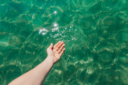 Woman hand reaches out to touch the sunlit, clear green water. Relaxing vacation activity concept.