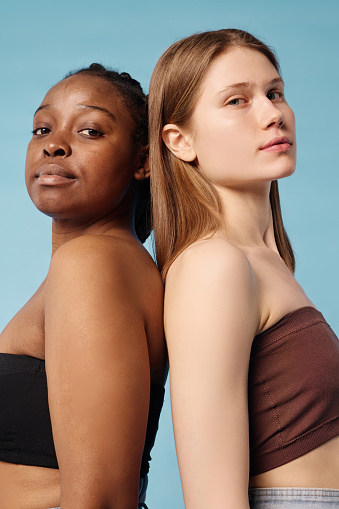 Vertical waist up studio portrait of two confident young Black and Caucasian women wearing bandeau tops standing back to back looking at camera