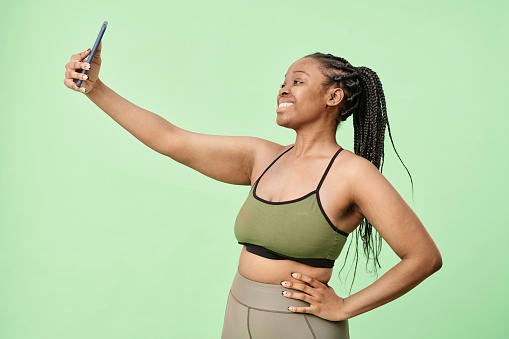 Medium portrait of modern young Black woman with braided hair wearing activewear standing in studio posing for selfie photos