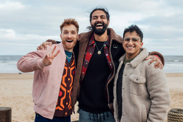 A shot of a small group of friends posing for a photo whilst spending time together at the beach in Tynemouth, North East England. They are smiling at the camera, wearing warm clothing. The male in the middle has his arms around his friends. The sea and beach are visible in the background.