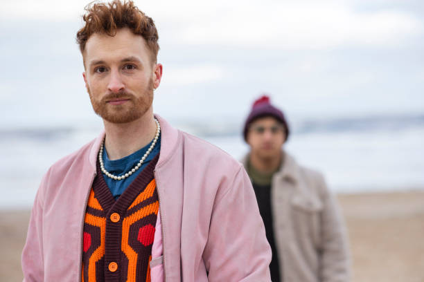 Fashionable Male at the Beach A waist-up portrait of an LGBTQIA male standing on the beach in Tynemouth, North East England. He is wearing layered colourful clothing and has a blank expression. It is a winter's day. Behind him is a blurred figure. queer man facing camera stock pictures, royalty-free photos & images