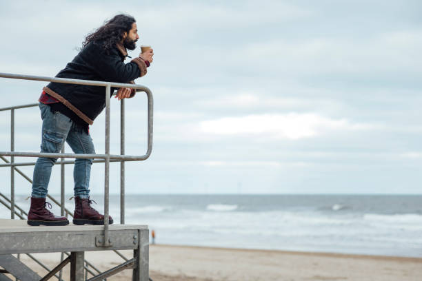 A side-profile full-length shot of an adult male leaning on a railing at the beach in Tynemouth, North East England. He is looking at the view whilst enjoying a takeout coffee. He has a beard and long hair, wearing warm clothing on a winter's day. The sea and beach are visible behind, out of focus.