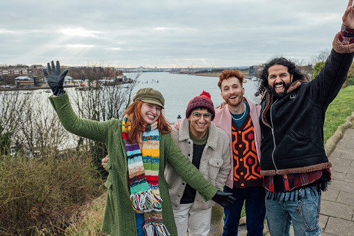 A shot of a small group of friends posing for a photo whilst spending time together at the beach in North Shields, North East England. They are smiling and two of them are raising their arms, all wearing warm clothing. It is a winter's day and the sea and quayside are visible in the background.