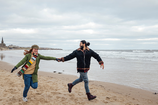 A full-length wide shot of two friends running along the beach together in Tynemouth, North East England. They are hand-in-hand, both smiling broadly. They are wearing warm clothing because it is a cold winter's day. In the background, waves roll towards the shore, and there is a headland beyond.