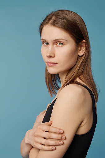 Vertical medium close-up studio portrait of young Caucasian woman standing with arms crossed looking at camera