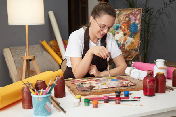 istock Artistic inspiration workshop. Creative student exhibition. Painter's artistic palette. Smiling Caucasian female artist wearing glasses and brown apron working on art project in her studio 2027403772