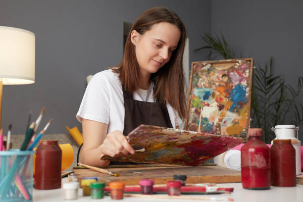 istock Artistic talent and imagination. Palette and brushes in use. Expressing creativity through paint. Caucasian female artist wearing brown apron working on art project in her studio 2027403778