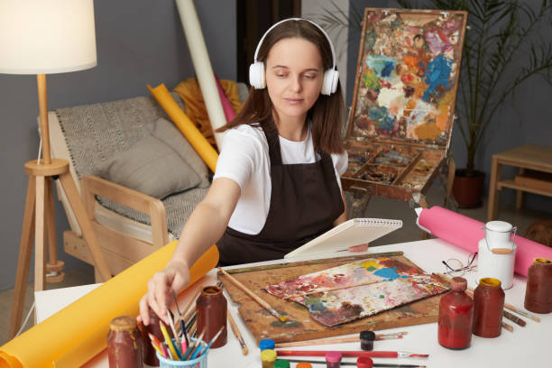 istock Workshop for creative minds. Showcasing artistic talent. Watercolor techniques and craft. Brown haired woman artist drawing with paints sitting in studio workshop and listening to music in headphones 2027403790