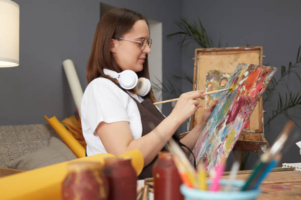istock Drawing inspiration from nature. Studio palette for vibrant paintings. Confident young Caucasian woman wearing glasses and brown apron painting with brush in artist studio 2027404302