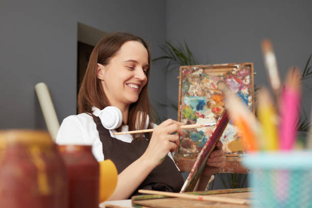 istock Professional artwork for an exhibition. Picture-perfect creative process. Smiling happy cheerful young Caucasian woman wearing brown apron painting with brush in artist studio 2027404327