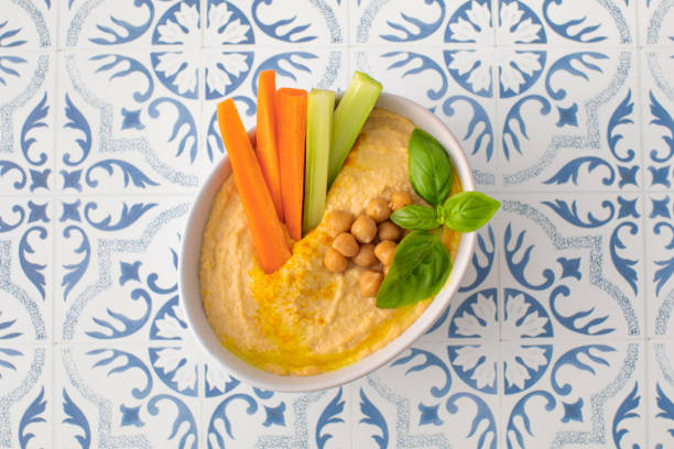 A bowl of hummus with carrots, cucumbers, and basil served on a table