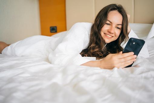 Smiling happy woman relaxing in bed and using smartphone