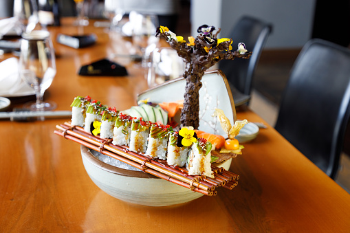 Sushi rolls lined up on bamboo place mat on top of bowl with crushed ice served on restaurant table, Japanese regional food