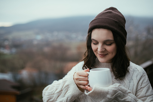 Young woman in white knitted sweater holding white cup, drinking hot tea, standing on a balcony of a cozy wooden house with mountains view.