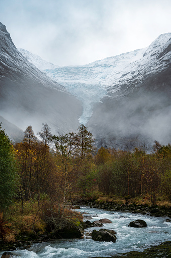 Norwegian mountain landscape, cloudy, with an autumn forest on a river, with the tongue of the Briksdalsbreen glacier in the background, over the snow-capped mountains.