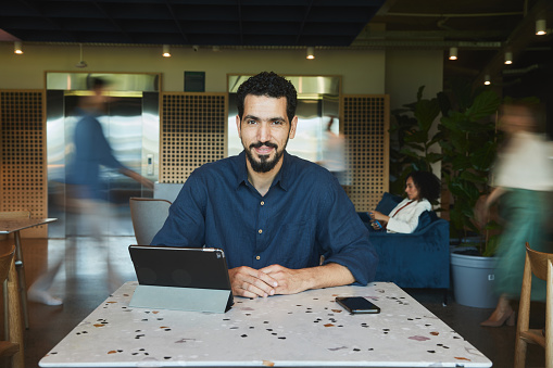 Portrait of a smiling young businessman working on a digital tablet at an office table with coworkers walking in blurred motion in the background