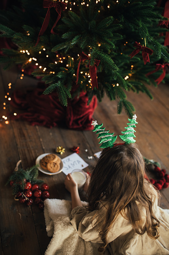 Cute 6 years old girl preparing milk and cookies for Santa under Christmas tree.