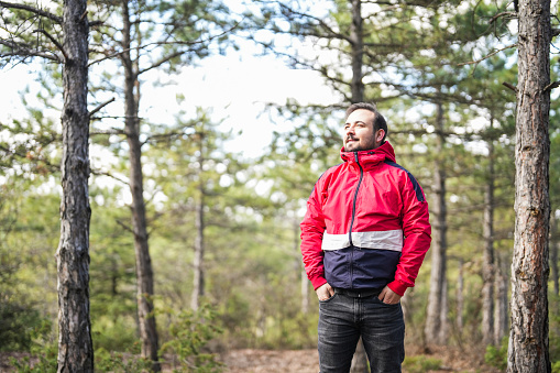 man in red coat standing in front of pine trees in the forest and looking at nature