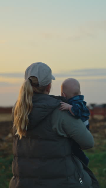 SLO MO Woman Farmer Carrying Baby Boy and Watching Tractor Plowing Field against Sky During Sunset