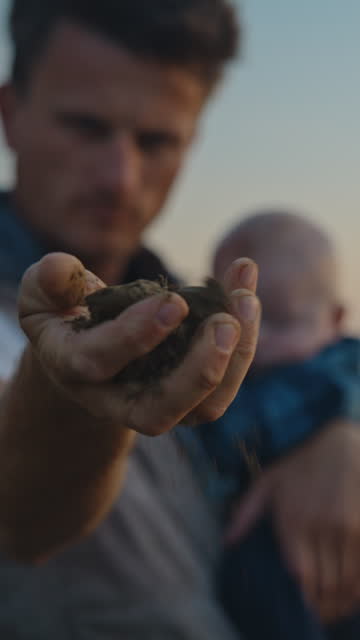 SLO MO Lockdown Shot of Male Agronomist Carrying Son While Checking Quality of Soil Before Sowing at Field During Sunset
