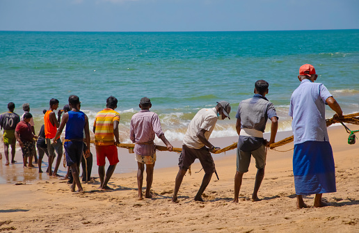 Unawatuna, Sri Lanka 09 february 2023 group of men in summer clothes of different colors helping fishermen to pull over large fishing net from ocean at beach of south Sri Lanka. hard team work.