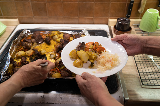 Close-up. One hand places the roast from the baking sheet onto a plate held by two hands. Background.