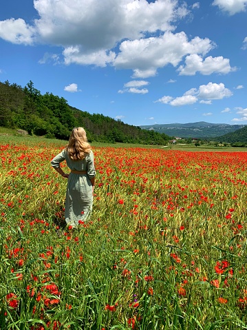 Woman in her 50s, dresswd in green dress standing in the middle of large poppy fields on a spring day with blue sky and white clouds