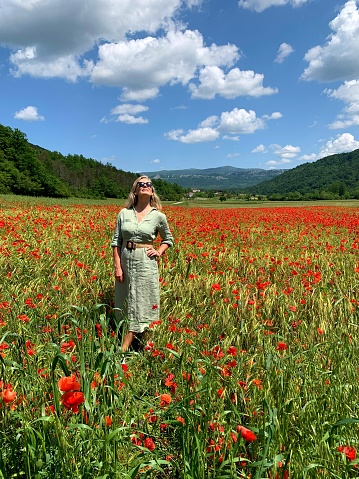 Woman in her 50s, dresswd in green dress standing in the middle of large poppy fields on a spring day with blue sky and white clouds