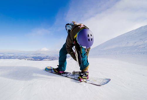 A person in winter gear snowboarding down a steep slope covered in fresh white snow. Girl preparing boots on her snowboard.