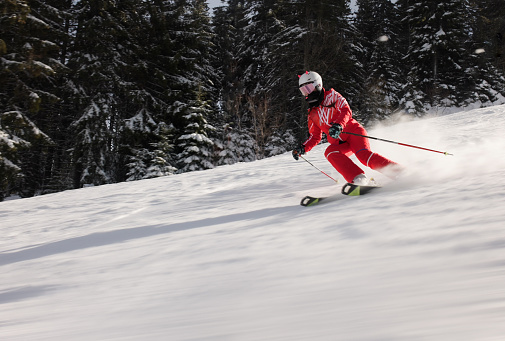 Female skier in a vibrant red ski suit skiing a snowy slope. Trees lining the run are frosted with snow. Cold environment typical of a winter sports setting. Crouched position for aerodynamics and a trail of powdered snow traces the rapid descent.